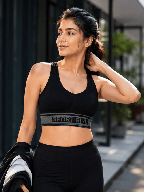 Woman in black sport bra with 'SPORT GIRL' band and matching leggings, holding jacket outdoors